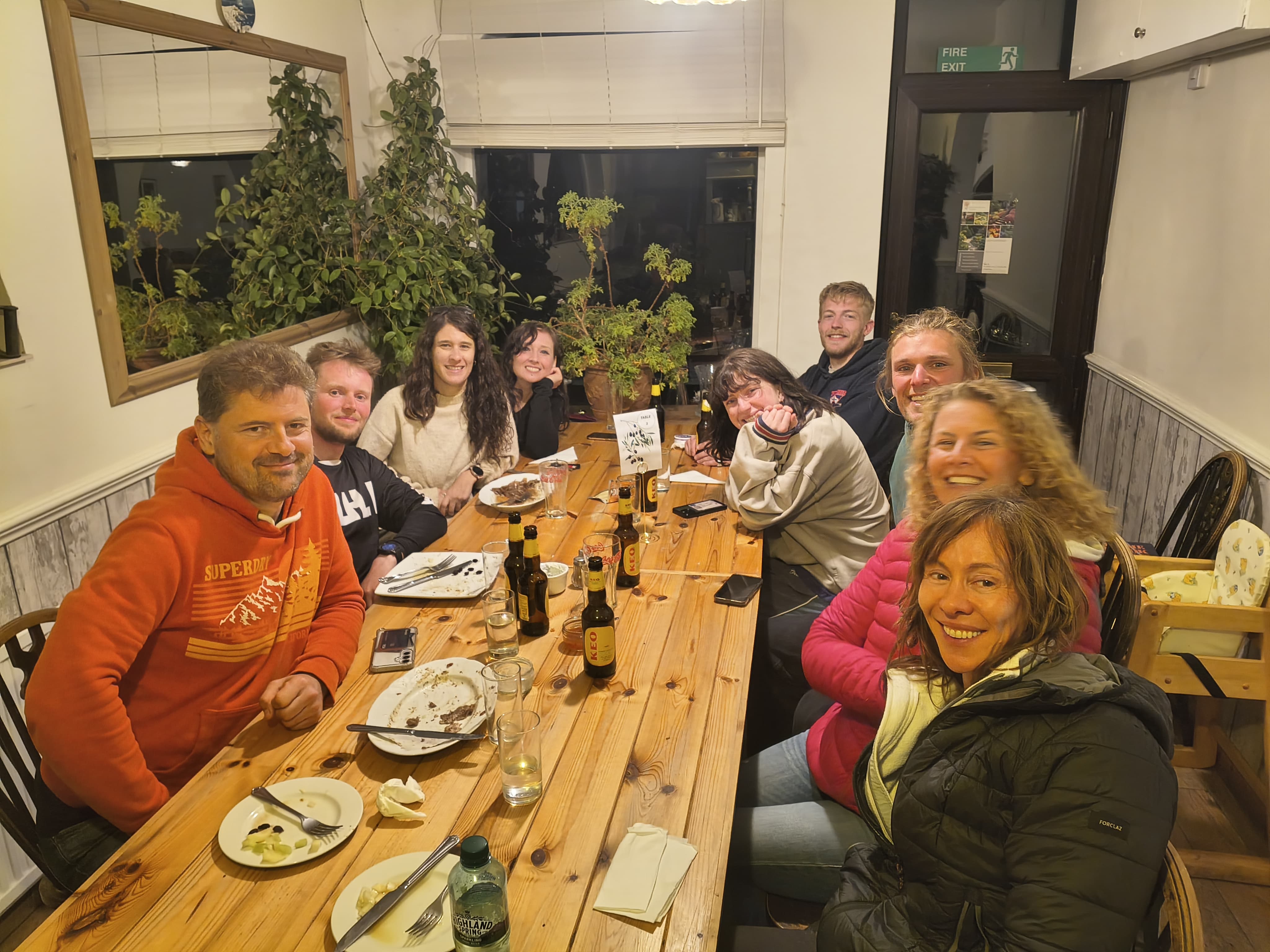 Smiling LKSA group around a long wooden table at a Greek restaurant, plates cleared, KEO bottles, indoor plants and warm lighting